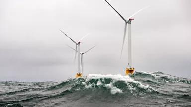 Three wind turbines stand in rough seas with a wave cresting in front of them.
