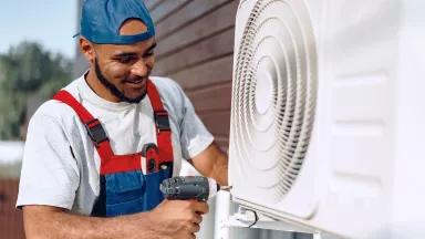 A worker holds a drill next to a white heat pump unit situated next to a building