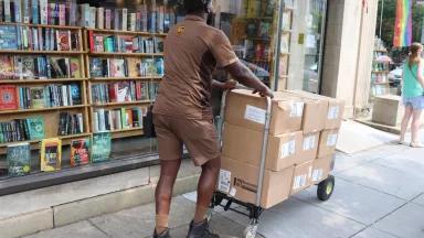 A UPS driver delivers packages with a wheeled dolly to a bookstore in Washington DC on a sunny summer day. He is wearing shorts and a short sleeved shirt, as is a pedestrian nearby.. 